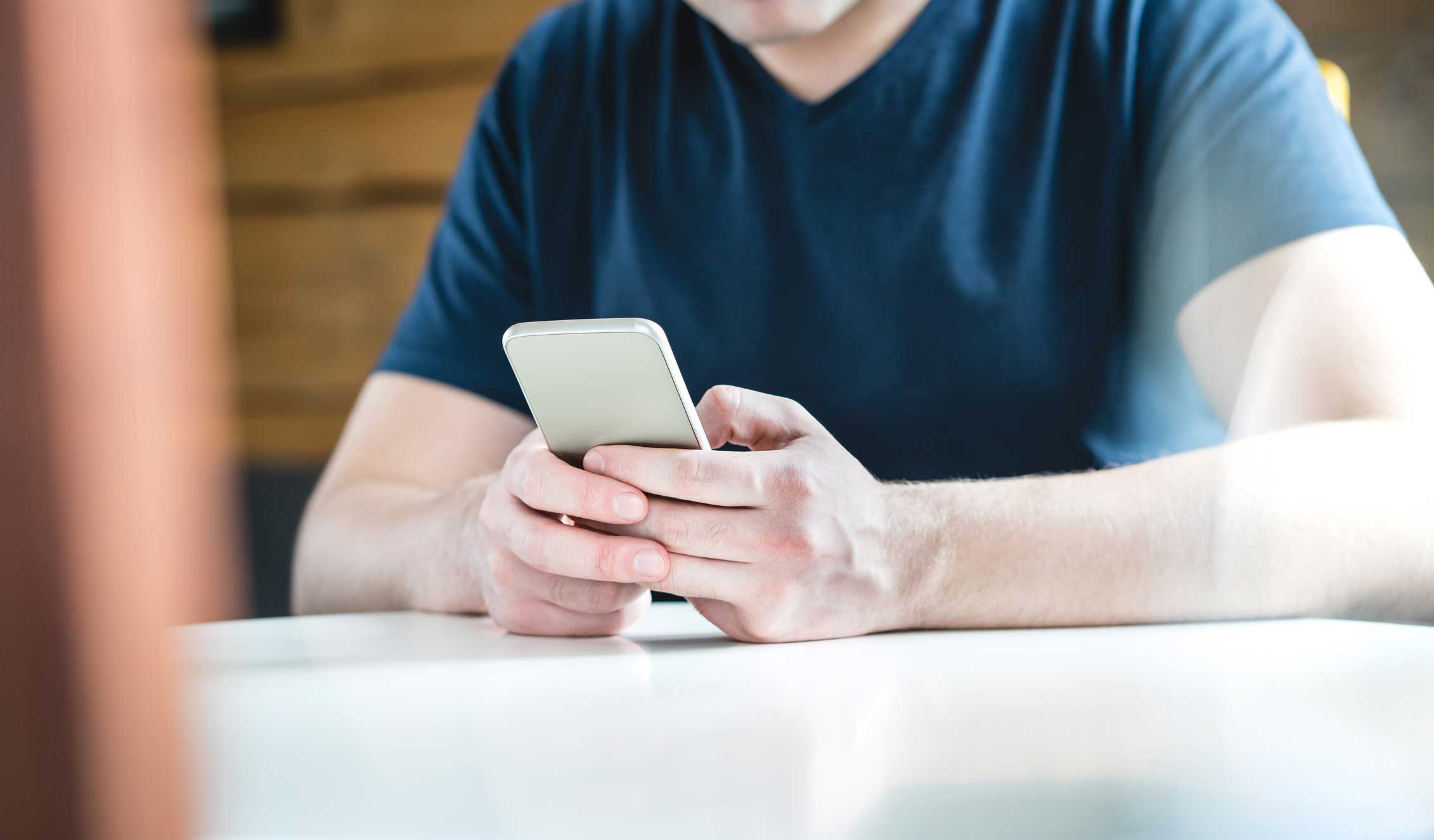 Young man texting with smartphone. Guy using mobile phone. Young man texting with smartphone. Guy using mobile phone against wooden wall and background. Cropped close up shot of hands holding cellphone on table.